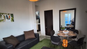 Ein Wohnzimmer mit einer schwarzen Ledersofa, einem runden Esstisch mit Stühlen, einem grünen Kunstgrasteppich und einer Pendelleuchte.