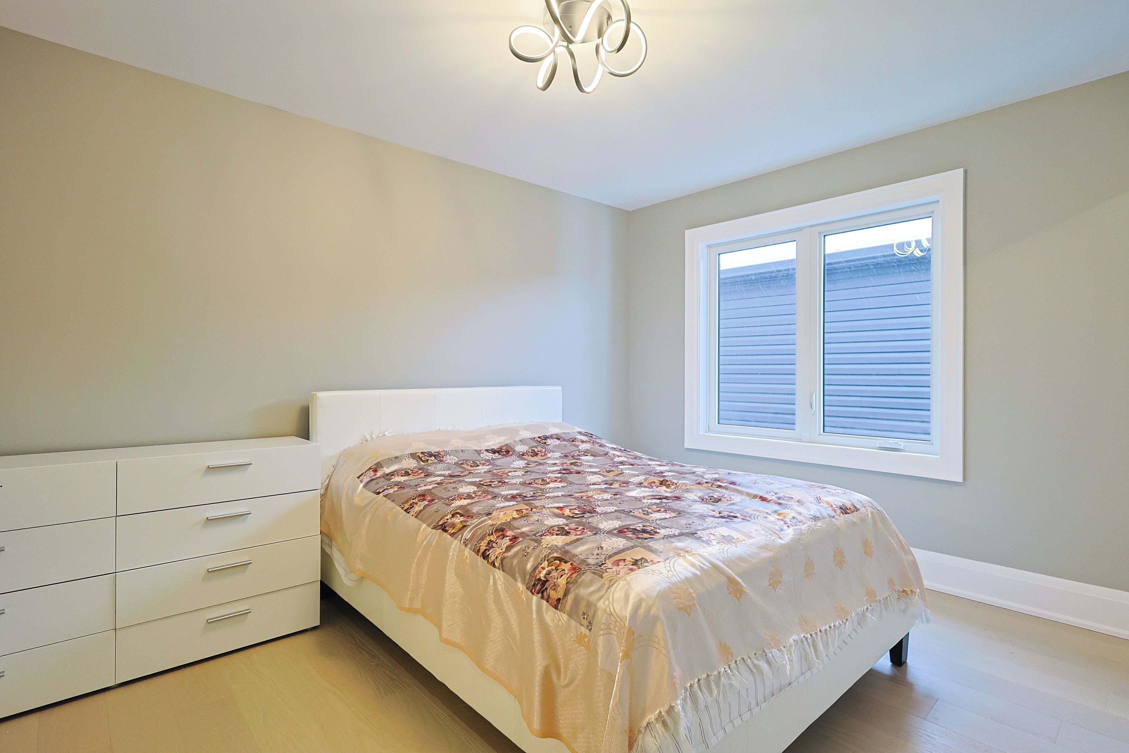 A bedroom with a bed, a dresser, a window with blinds, and a ceiling light fixture.