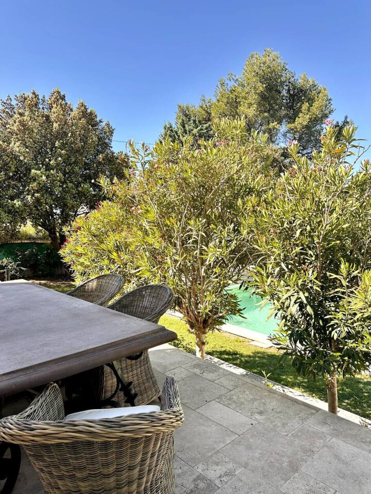 An outdoor patio with a table and wicker chairs, surrounded by trees and a swimming pool.