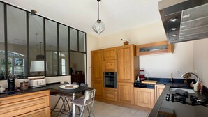 A modern kitchen with wooden cabinets, a dining table with chairs, a built-in oven, and a gas stove. - Les Lauriers Roses, air-conditioned villa with garden and swimming pool. (L'Isle-sur-la-Sorgue)