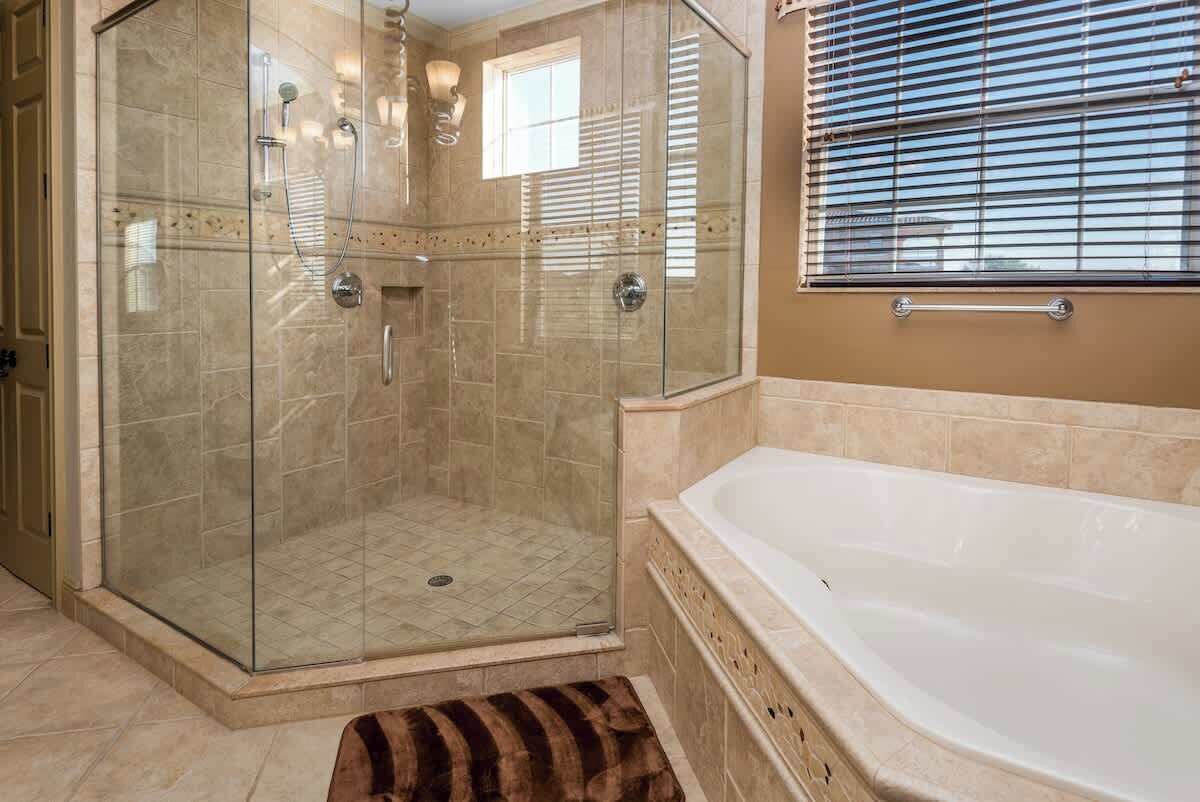Een douche met glazen wanden en een inloopdouche, een wit bad en een raam met zonwering.
