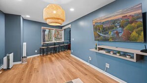 A modern living room with a large TV, a dining area with bar stools, a hanging light fixture, and a cityscape painting on the wall. - The Lazy Lodge: Peaceful Retreat 15 mins from Downtown Greenville (Easley)