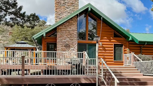 A log cabin with a stone chimney, large windows, and a wooden deck with a fence.