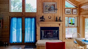 A wooden cabin interior with a fireplace, a dining area, and a large window with curtains.