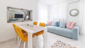 Een moderne woonkamer met een witte eettafel, gele stoelen, een lichtblauwe bank met roze en blauwe kussens, een ingelijste afbeelding en een spiegel aan de muur.