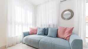 Een lichtblauwe bank met roze kussens in een lichte kamer met witte gordijnen en een ronde spiegel aan de muur.