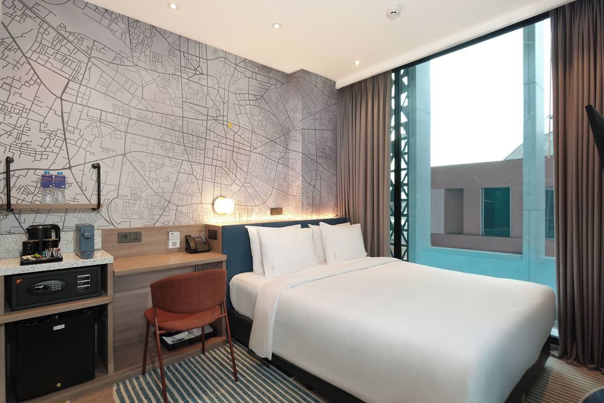 A hotel room with a large bed, a desk with a computer, a chair, a map on the wall, and a window with curtains.