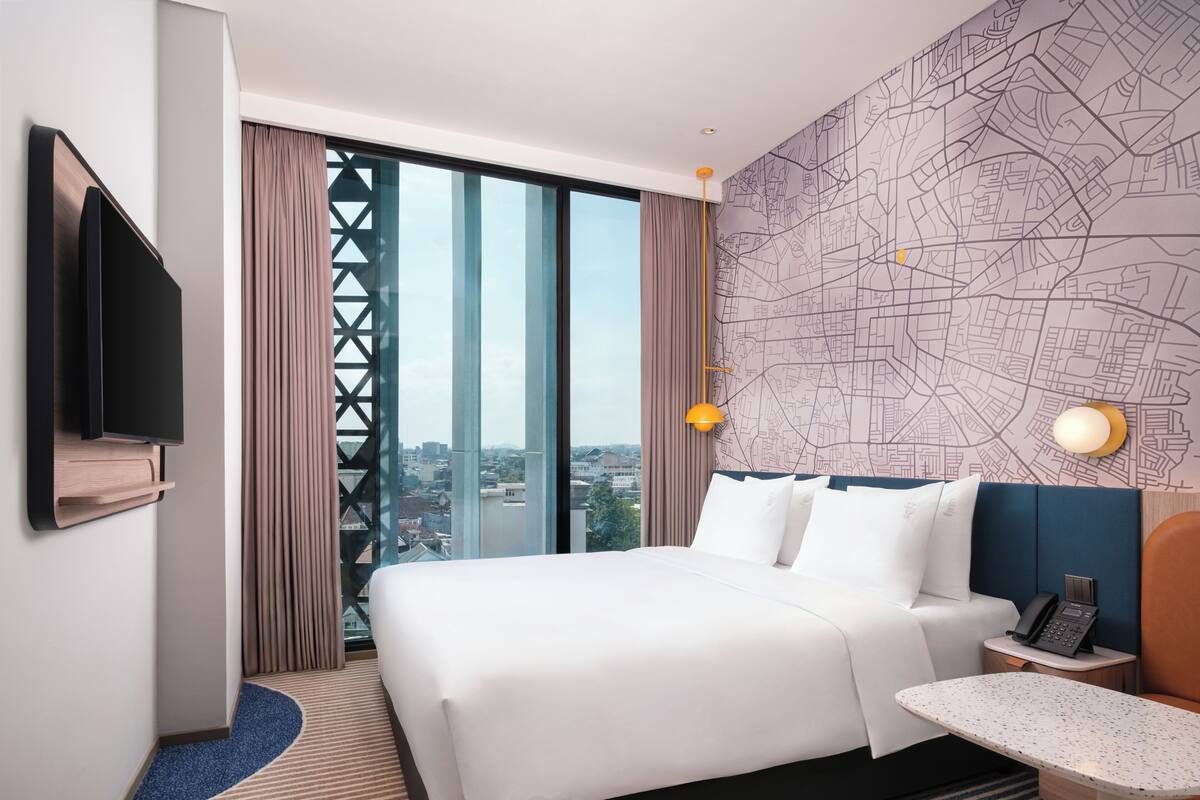 A hotel room with a large bed, a desk, a phone, a TV, and a cityscape view.