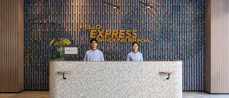 A reception desk with two staff members, a patterned wall, and a sign reading 'HELLO EXPRESS BANDUNG BRAGA'.