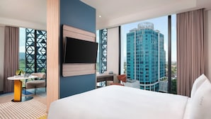 Ein Hotelzimmer mit Bett, Fernseher, Schreibtisch und Stuhl. Ein großes Fenster bietet Blick auf ein hohes Gebäude.