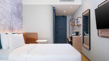 Una habitación de hotel con cama, una mesita, una silla, un espejo, un televisor y una puerta que da al baño.