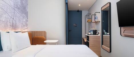 Una habitación de hotel con cama, una mesita, una silla, un espejo, un televisor y una puerta que da al baño.