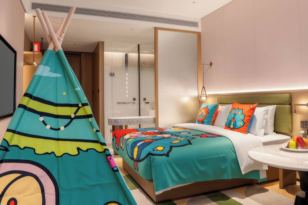 Quarto de hotel moderno com cama, mesinha, cadeira, luminária pendente e uma janela grande.