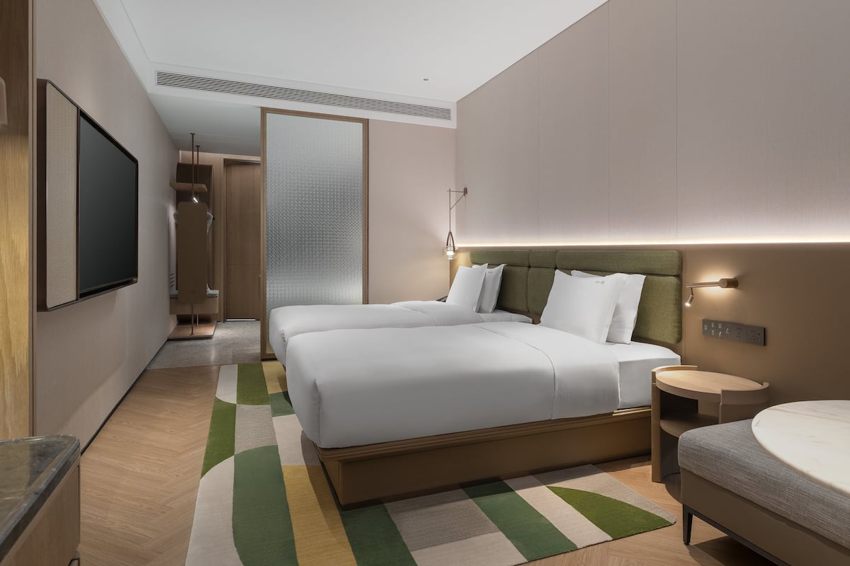 Quarto de hotel moderno com uma cama grande, mesas de cabeceira, televisão e banheiro com pia e espelho.