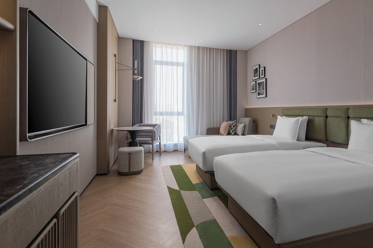 Quarto de hotel com duas camas, televisão, mesa pequena e janela com cortinas.