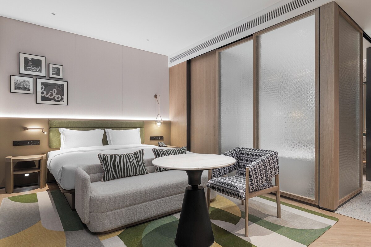 Moderni hotellihuone, jossa on sänky, yöpöytä, lamppu, sohva, tuoli ja suuri liukuovi.