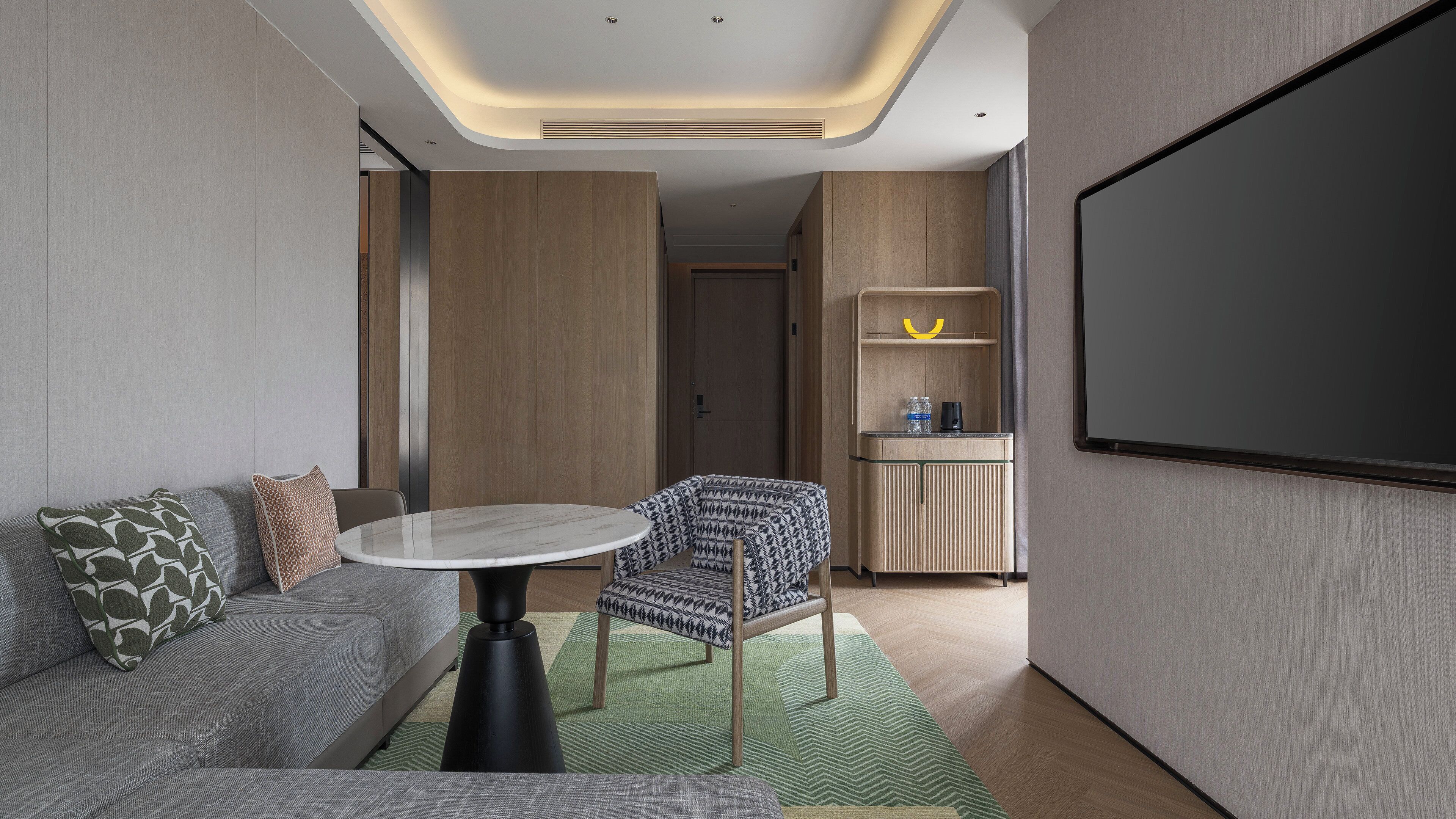 A modern hotel room with a sofa, a small round table, a patterned armchair, a projector screen, and a kitchenette.