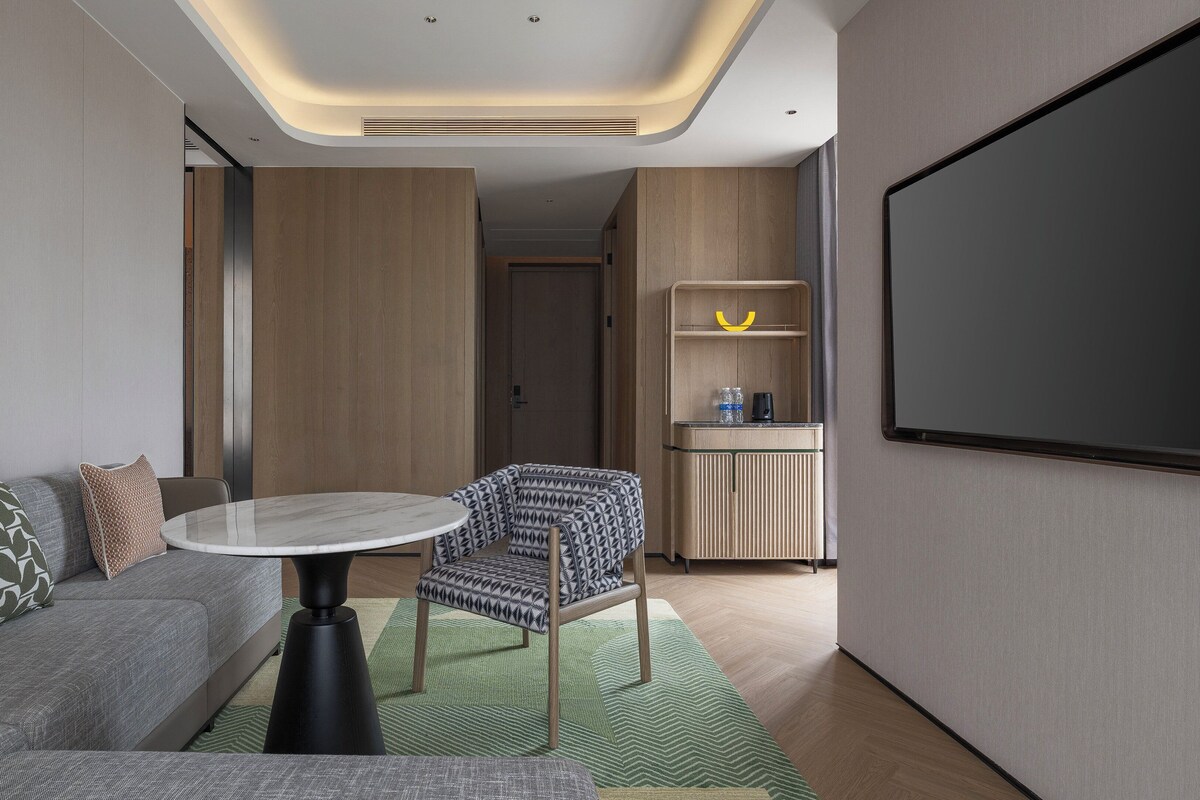 Moderni hotellihuone, jossa on sohva, pieni pyöreä pöytä, kuviollinen nojatuoli, projektorikangas ja pieni keittiö.