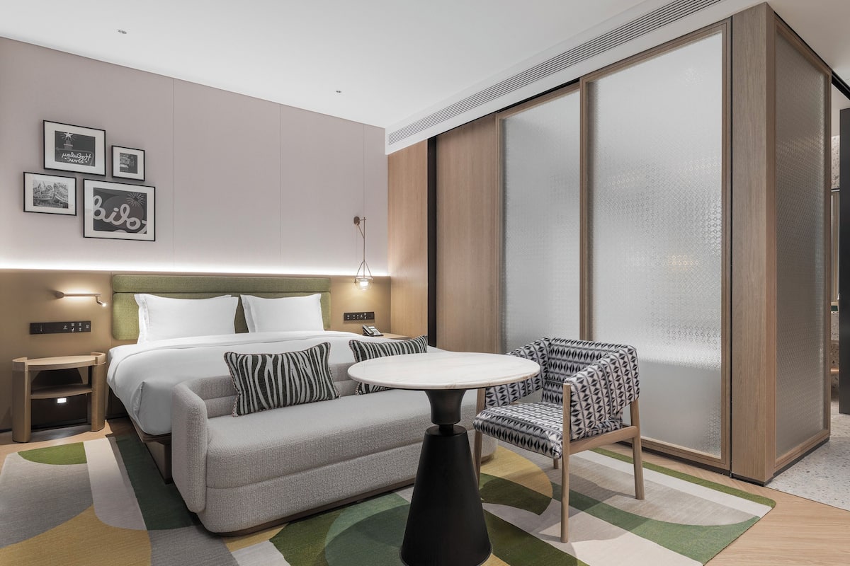 Quarto de hotel moderno com cama, mesa de cabeceira, abajur, sofá, poltrona e uma grande porta deslizante.