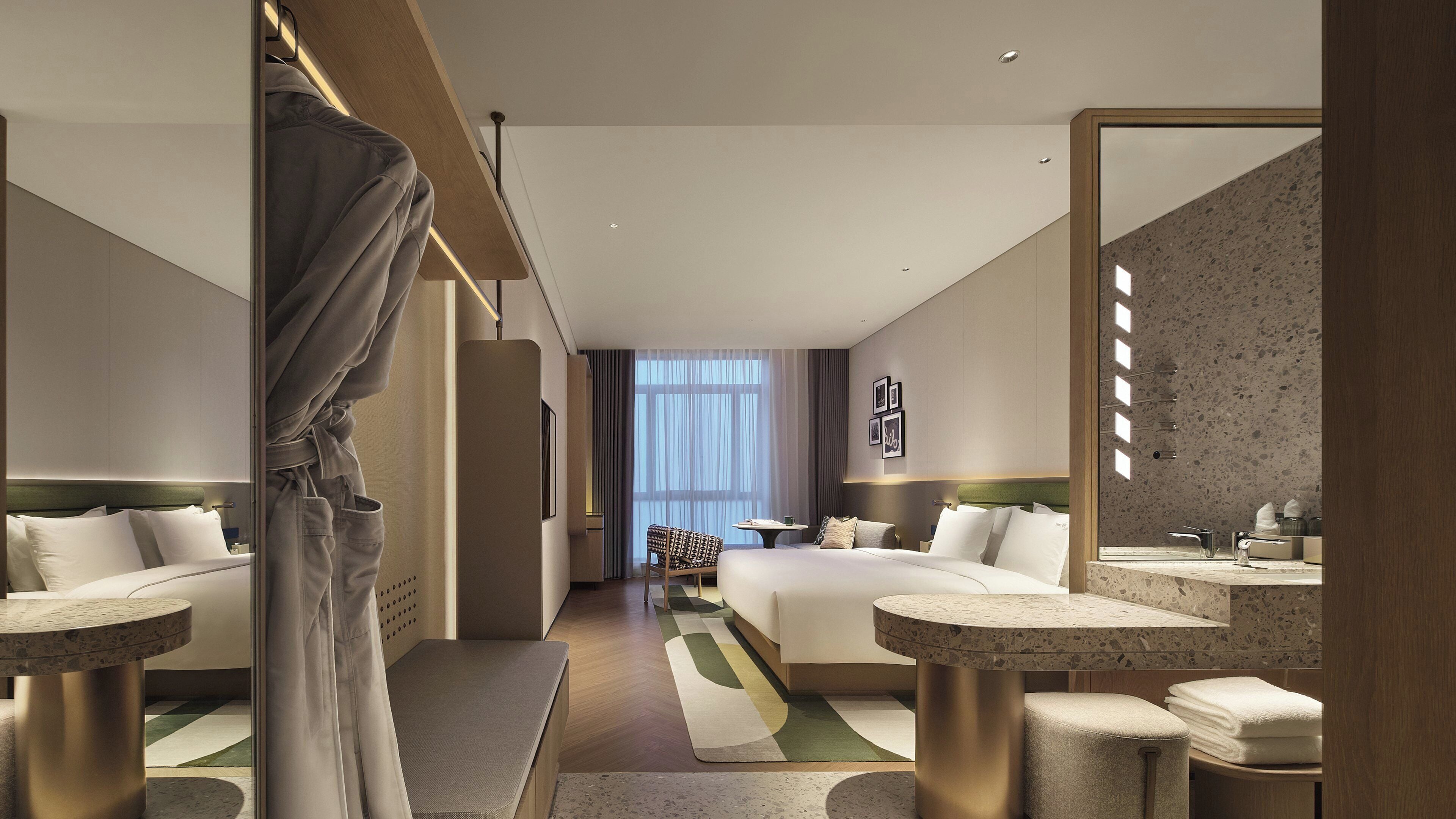 Ein Hotelzimmer mit einem Bett, zwei Nachttischen, einem Sessel und einem Spiegel, mit Blick auf den Flur.