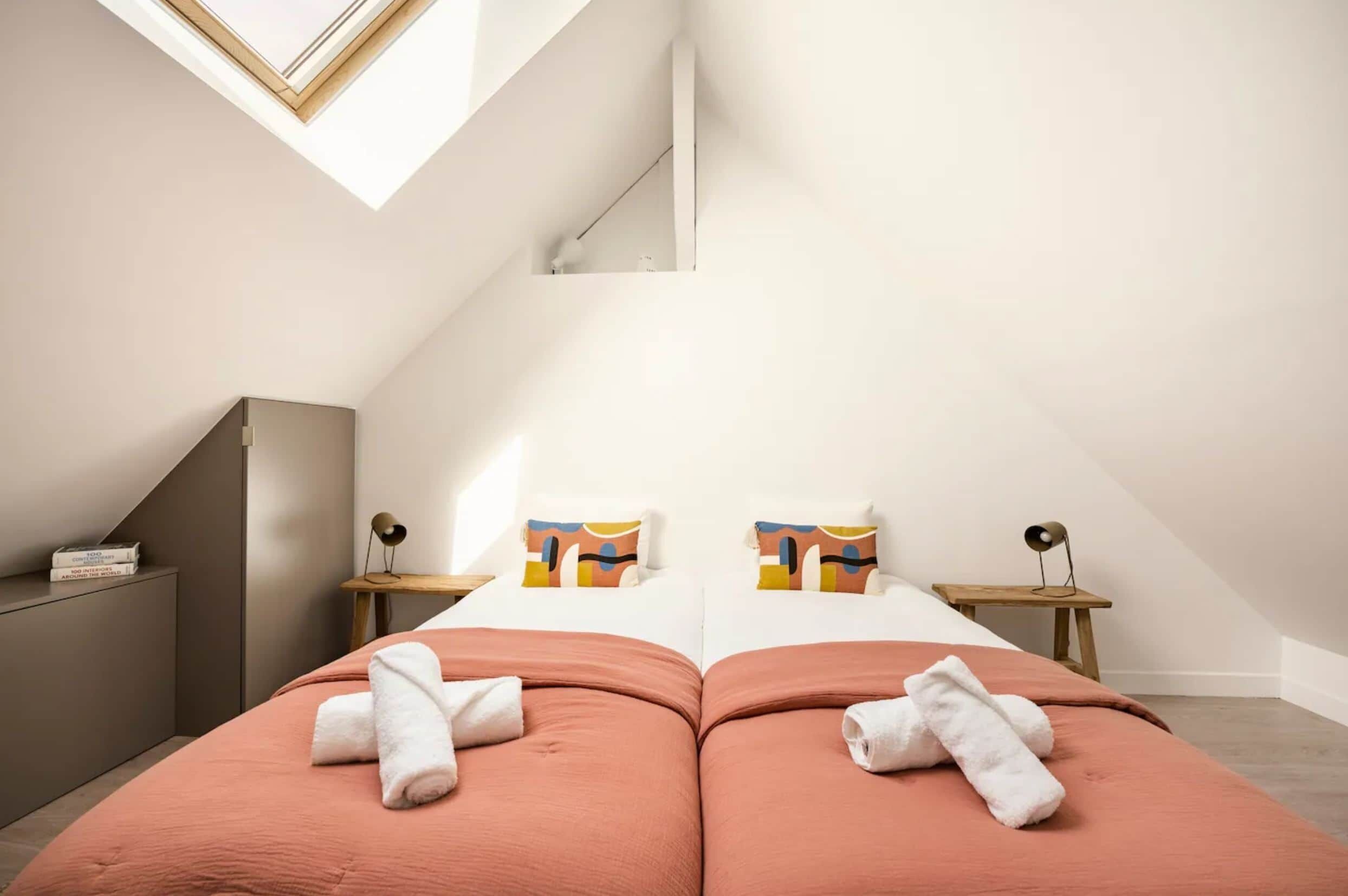 Een moderne slaapkamer met dakraam, twee bedden met handdoeken, een klein tafeltje en een boekenkast.