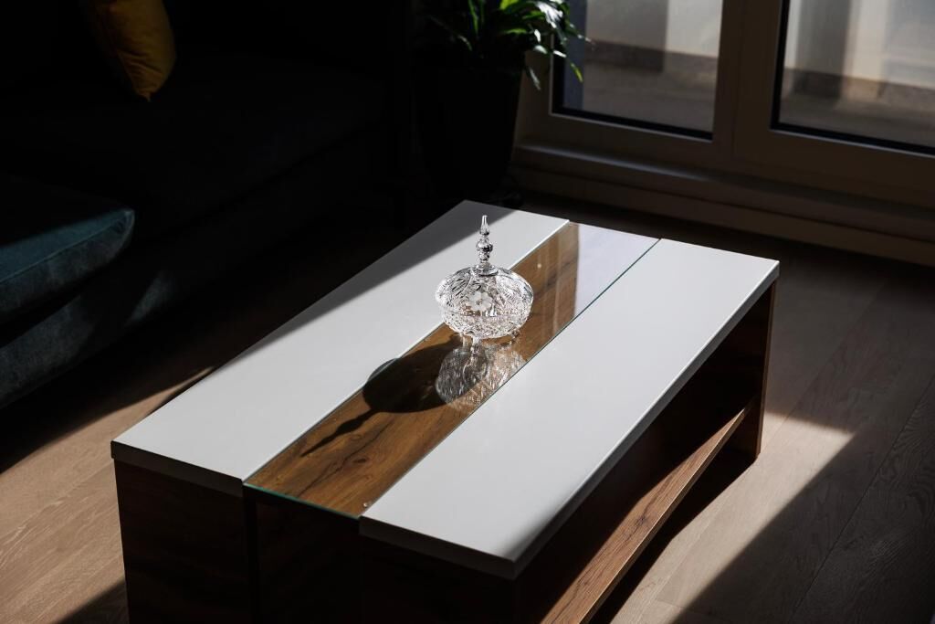 A modern living room with a glass coffee table, a wooden accent, and a view through a window.