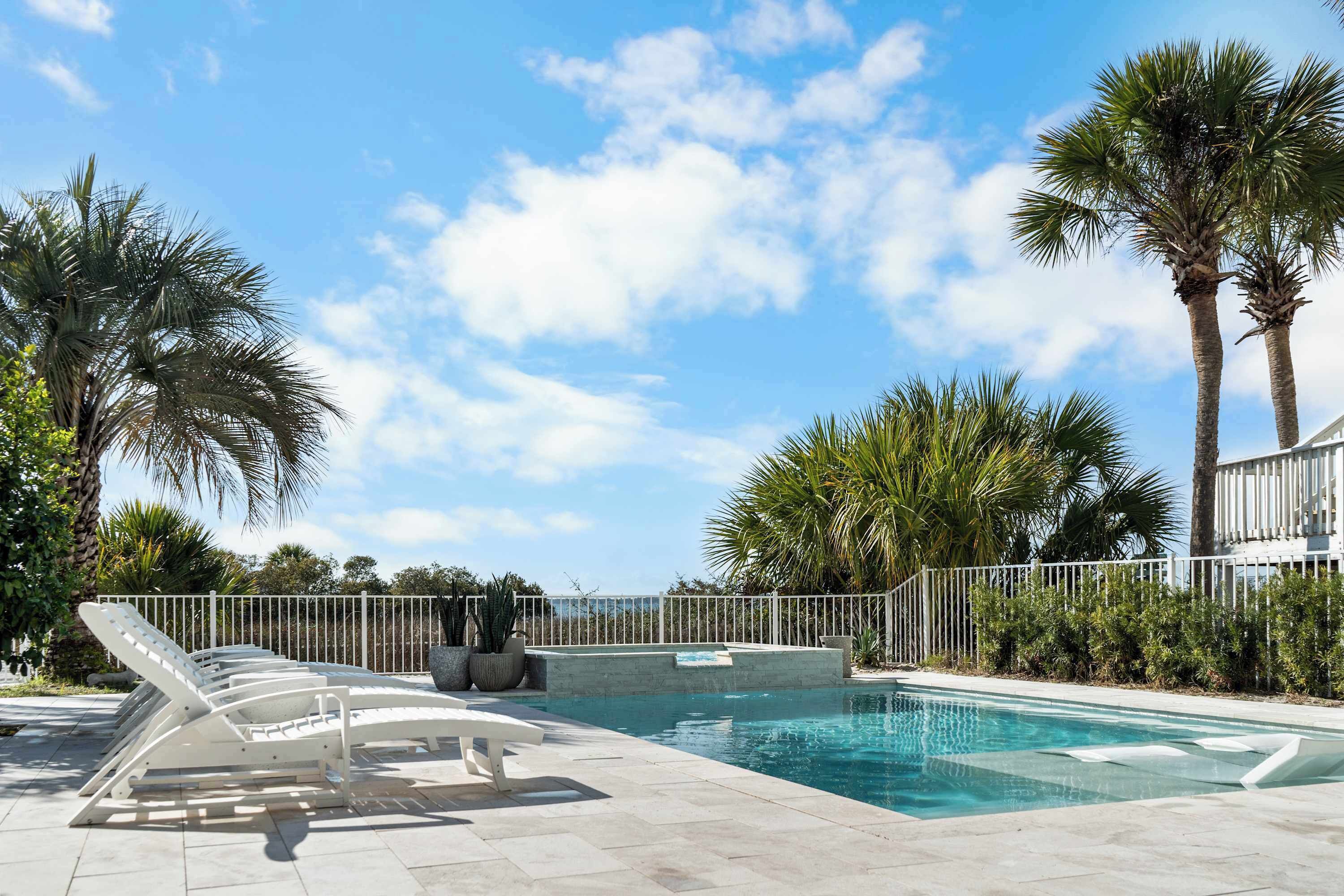 A swimming pool with lounge chairs, palm trees, and a white fence.