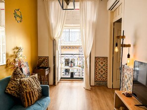 A room with a blue sofa, a wooden side table, a decorative plant, and a view through a window with white curtains. - A cozy little house in the center of Lisbon, featuring well-preserved historic architecture.   (Lisboa)