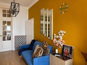 Uma sala de estar com um sofá azul, uma mesinha de madeira, um vaso com flores, um quadro emoldurado e um espelho na parede.