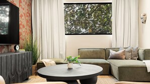 Un salon moderne avec un canapé d’angle, une table basse ronde, un téléviseur à écran plat et une grande fenêtre donnant sur la forêt.