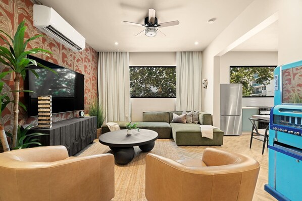 A modern living room with a flat-screen TV, a ceiling fan, a dining area, and a kitchen with a blue oven. - Condado 5BR Apt| Walk to Beach & Restaurants (San Juan)