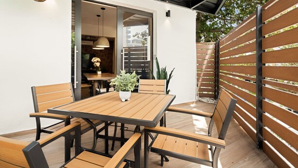 Une terrasse extérieure avec une table et des chaises en bois, une plante suspendue et une vue sur l’intérieur par une porte ouverte.