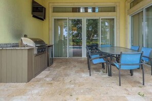 Une terrasse extérieure équipée d’un barbecue, d’une table avec des chaises et d’une porte-fenêtre coulissante donnant sur un jardin.
