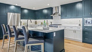 Een moderne keuken met donkerblauwe kastjes, een centraal eiland met barstoelen, roestvrijstalen apparatuur en een witte achterwand.