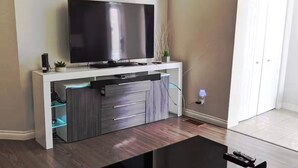 Een moderne woonkamer met een flatscreen televisie op een witte tv-meubel, een salontafel met glazen blad, en een vloer van donker hout.