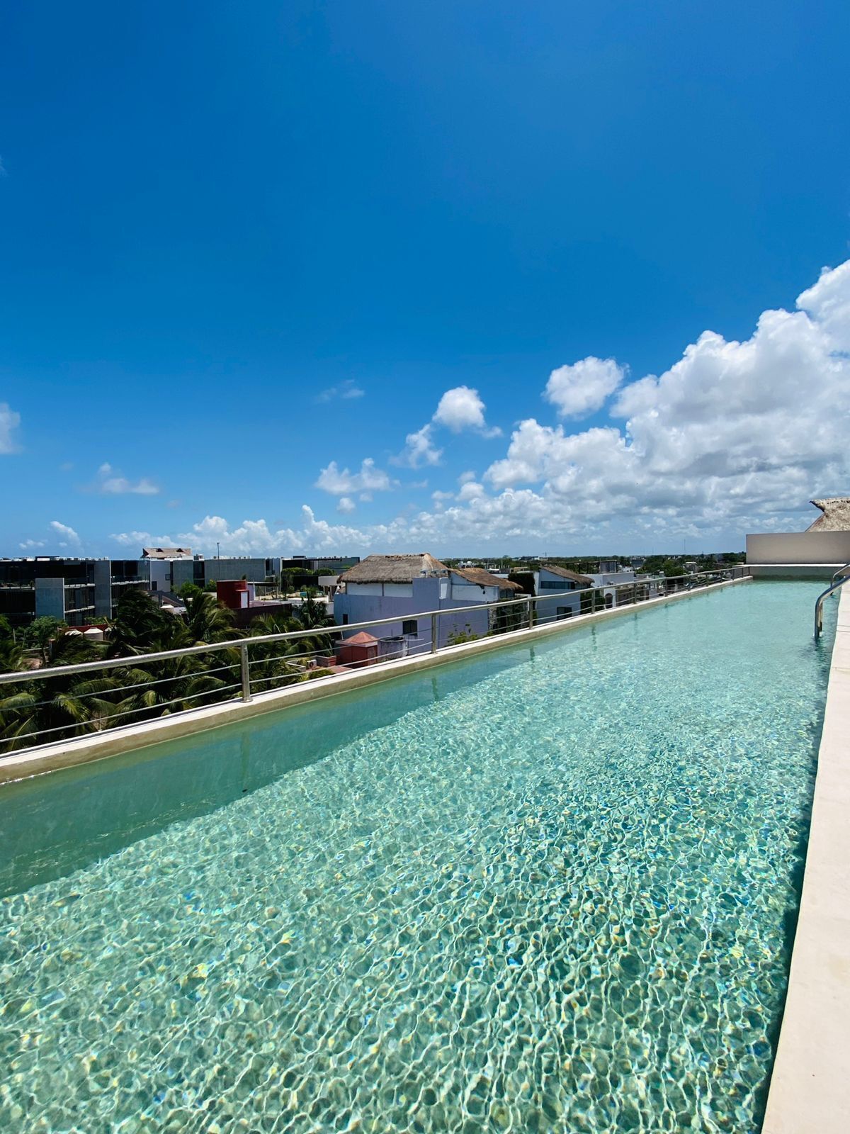 A clear pool with a metal railing, surrounded by buildings and palm trees under a blue sky.