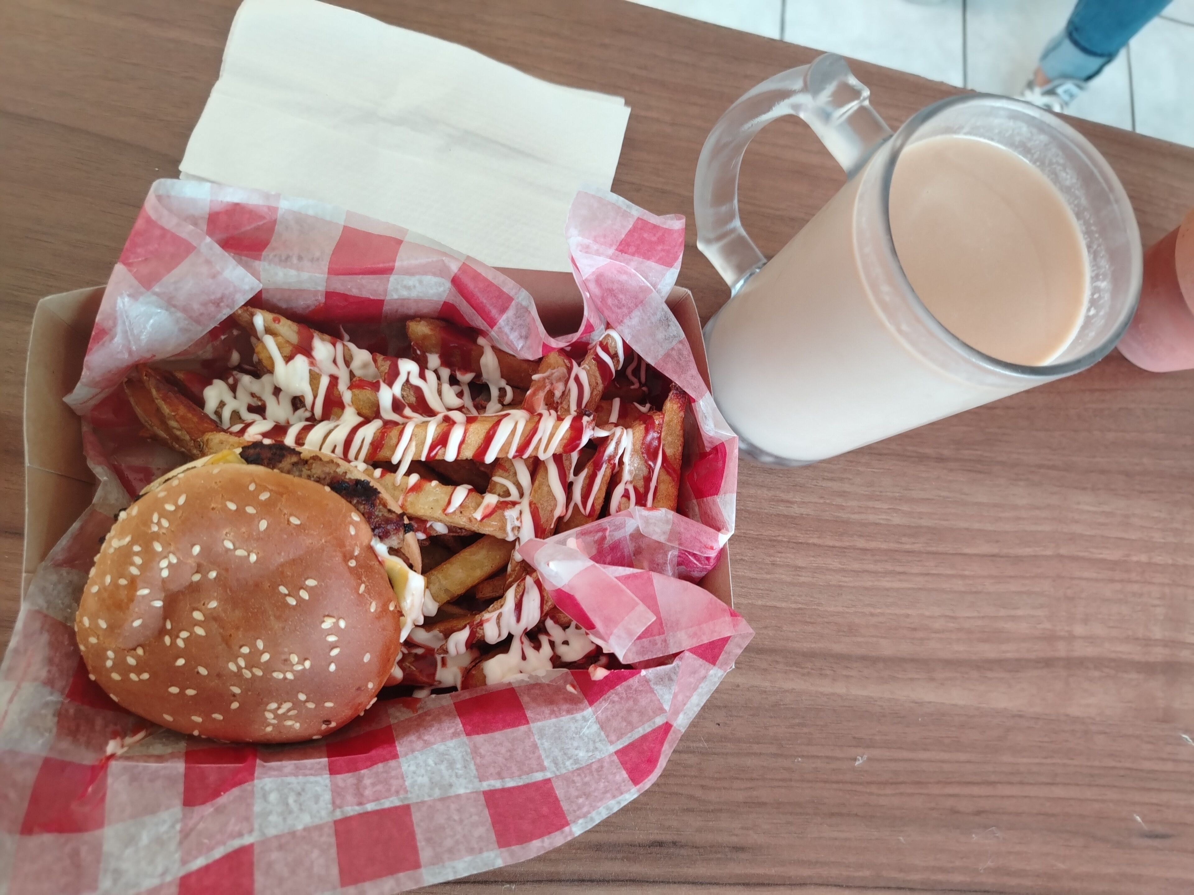 A meal consisting of a burger and fries, served on a checkered paper, with a glass of a light-colored beverage beside it.