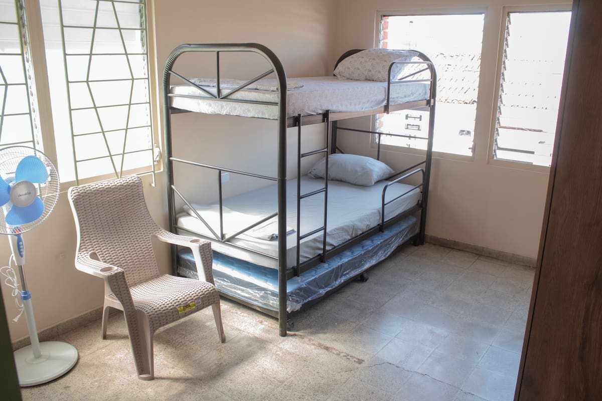 A bunk bed with a chair and a standing fan in a room with windows.
