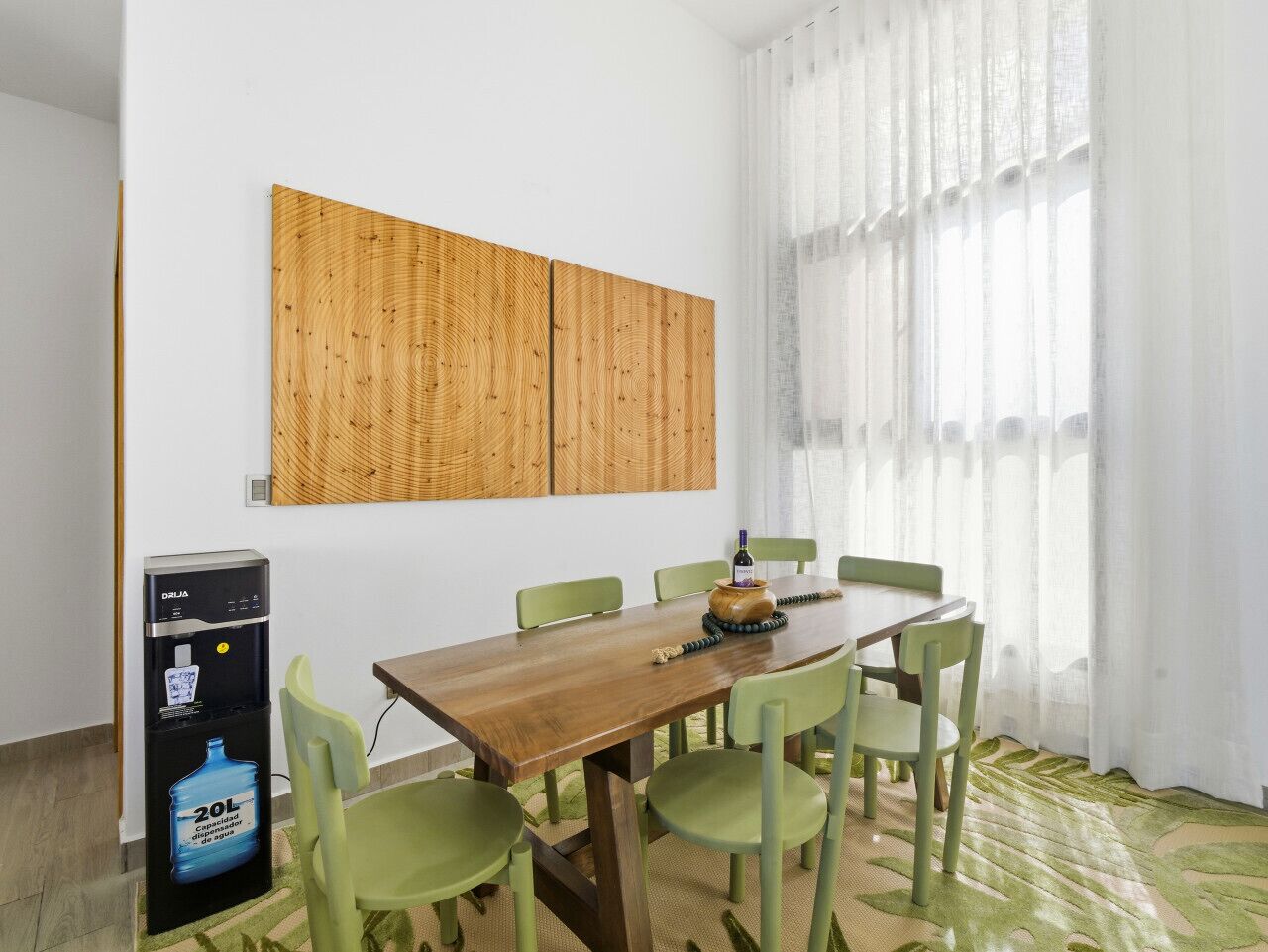 Un espace repas comprenant une table en bois, des chaises vertes, un distributeur d’eau et une grande fenêtre ornée de rideaux légers.