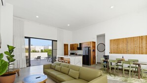 A modern living room with a sofa, a dining table with chairs, a kitchen area, and a large window.