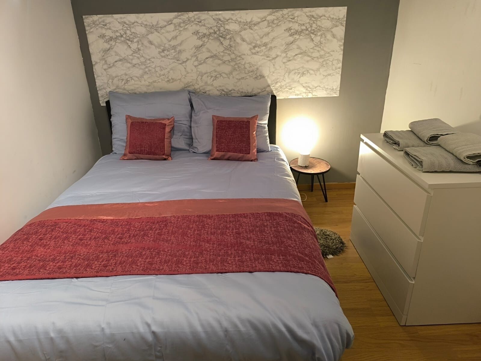 Un lit avec une tête de lit en marbre, deux oreillers, une couverture rouge, une lampe de chevet et une pile de serviettes sur une commode.
