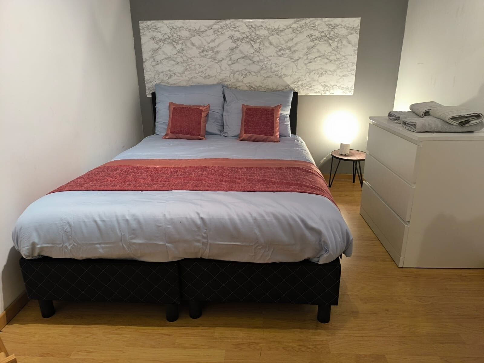 A neatly made bed with a headboard, a small bedside table with a lamp, and a dresser.