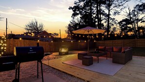 Ein Garten mit einer Holzterrasse, einem Grill, Gartenmöbeln und Lichterketten.