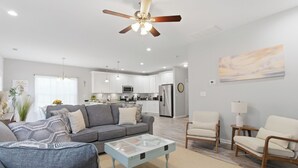 Ein modernes Wohnzimmer mit Sofa, Couchtisch und Sesseln, im Hintergrund eine Küche und ein Deckenventilator.
