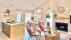 Una cocina moderna con una isla central, comedor con sillas de mimbre, chimenea y sala de estar con televisor.
