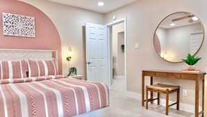 Un dormitorio con cama, mesita de noche, espejo y una puerta que da a otra habitación.