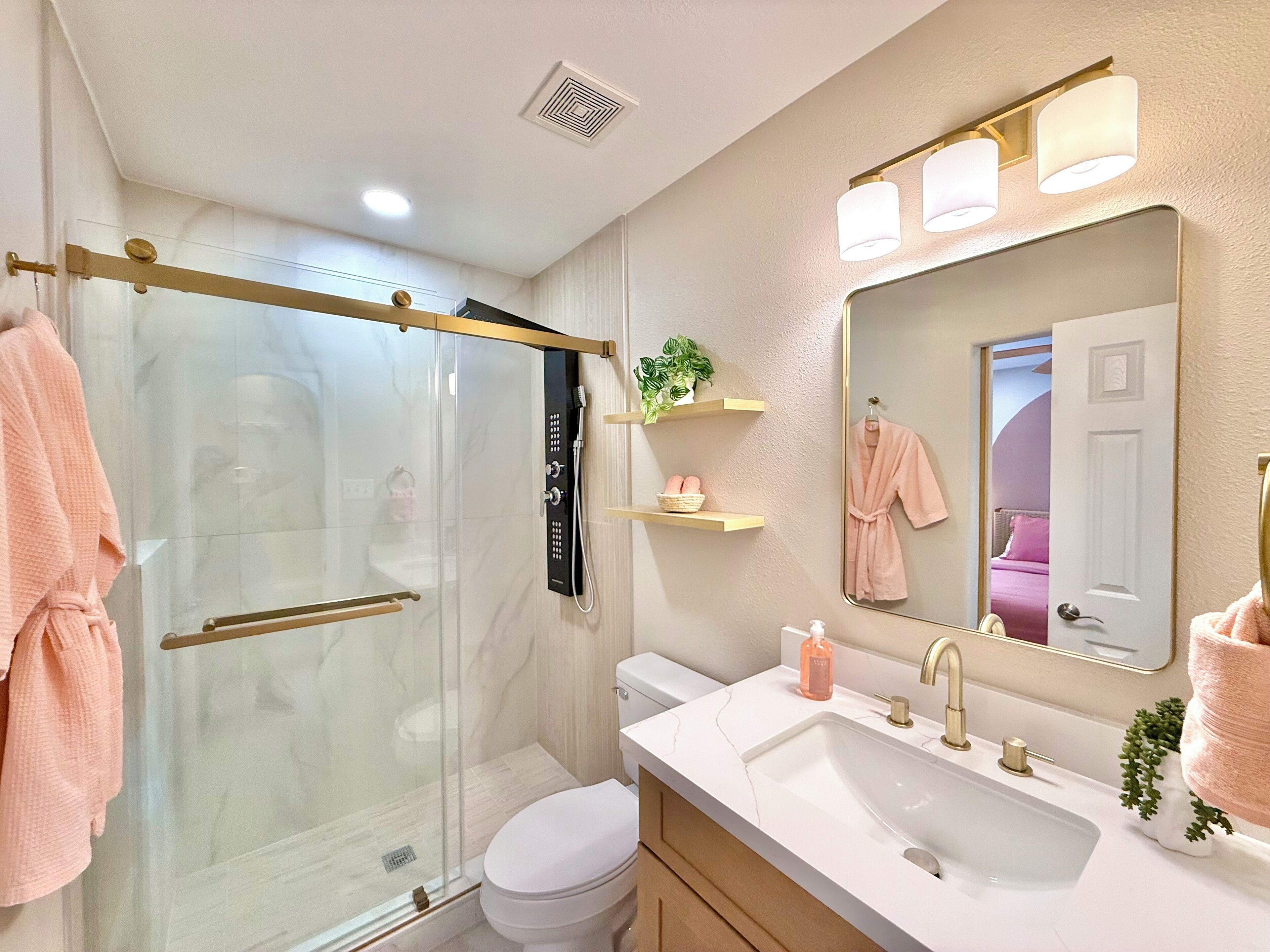 A bathroom with a glass-enclosed shower, a white sink with gold fixtures, a large mirror, and a pink robe hanging on the wall.