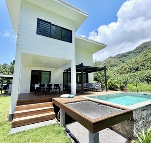 Et moderne hus i to etager med pool, udendørs siddepladser og udsigt til en frodig, grøn bakke.