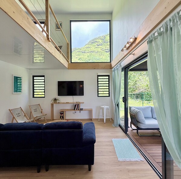 A modern living room with a large TV, wooden beams, a sofa, a small table, and a view of greenery.