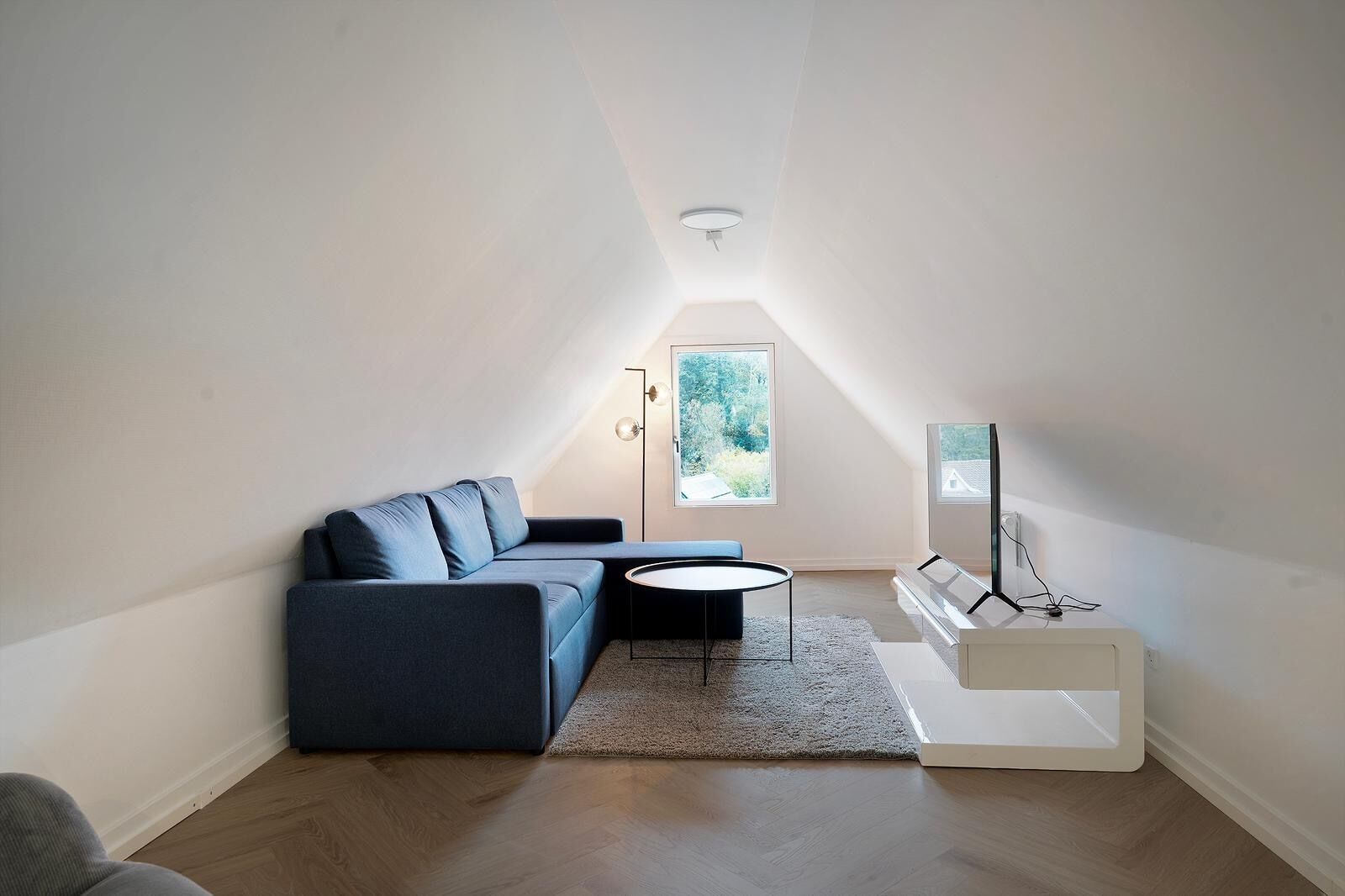 A modern loft bedroom with a sloped ceiling, a built-in TV stand, a sofa, and a small round table.